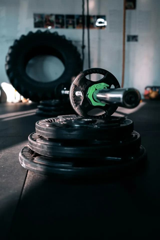 A stack of weight plates and a barbell on a gym floor, with a large tractor tyre behind