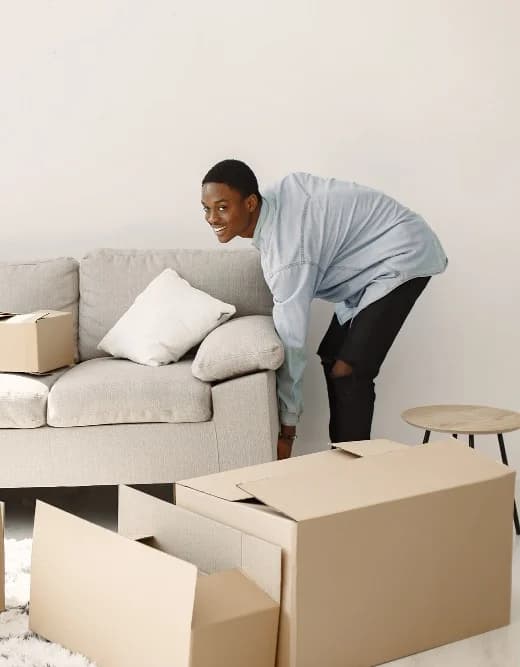 A smiling man lifting a light grey sofa, surrounded by moving boxes