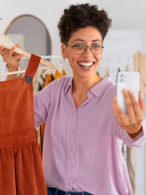 A smiling woman holding up a rust-coloured pinafore dress on a hanger while taking a photo of it with her phone