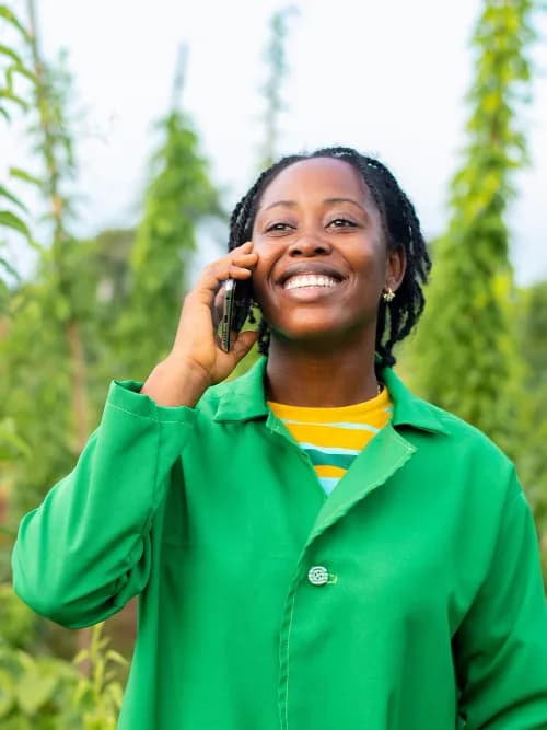 A smiling woman in a green jacket talking on her phone among tall green plants