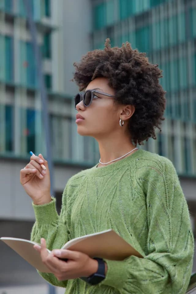 A student in sunglasses and a green sweater holding a notebook and pen outside a glass building